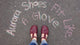 A customer is wearing a pair of Burgundy Middle T-Strap shoes by Aurora Shoe Co.    The photo is taken from the customer's perspective, looking down at her new burgundy handmade T-straps.   In Chalk, the words "Aurora Shoes Fit Like A Glove" is written on a blacktop surface, framing her new shoes in the photo.